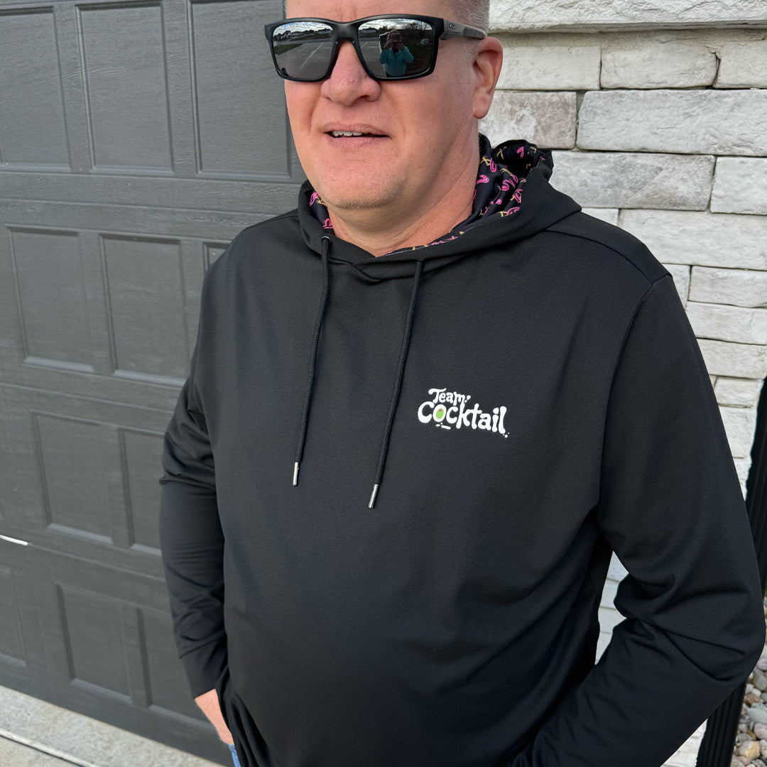 Man wearing a black hoodie with a logo and text, standing against a gray wall.