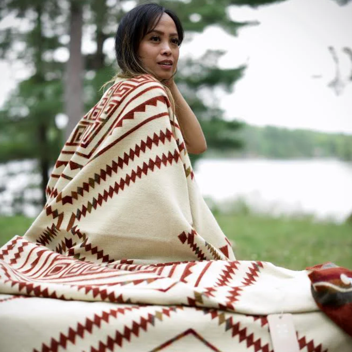 Woman sitting outdoors wrapped in a patterned blanket with a blurred natural background