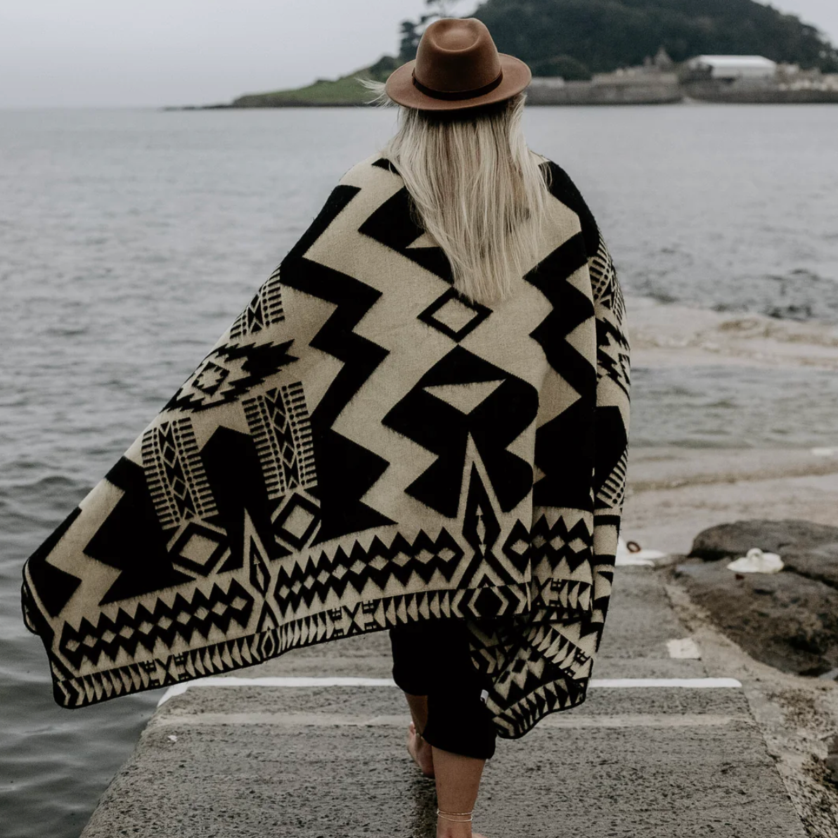 Person wearing a black and white patterned poncho and brown hat by a body of water.