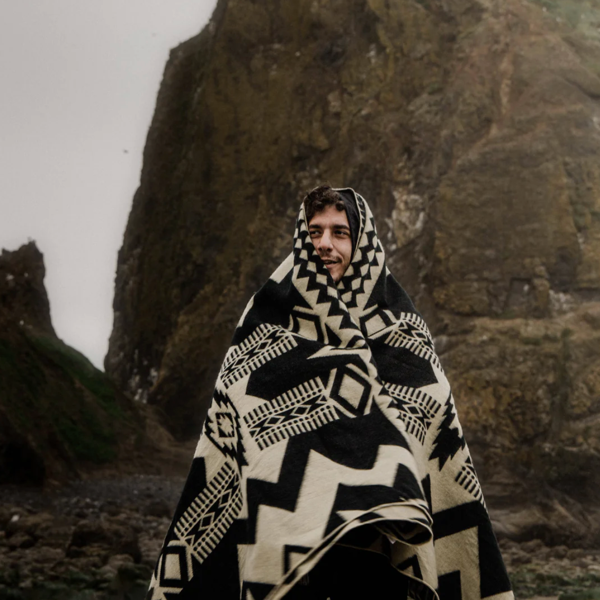 Person wrapped in a black and white patterned blanket standing against a rocky background