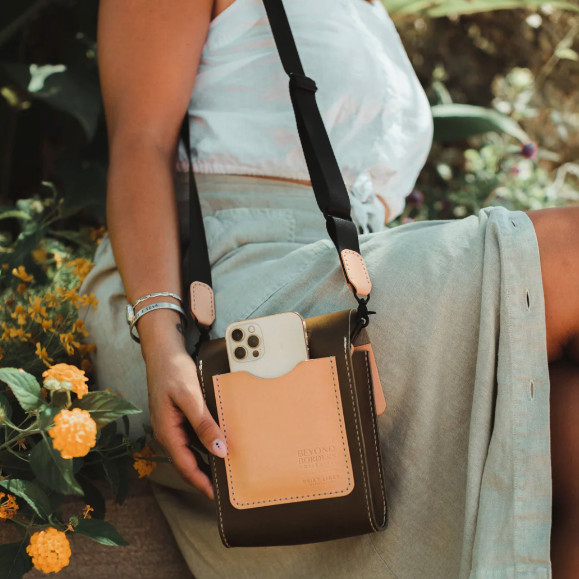Person holding a brown crossbody bag with a phone pocket, surrounded by flowers.
