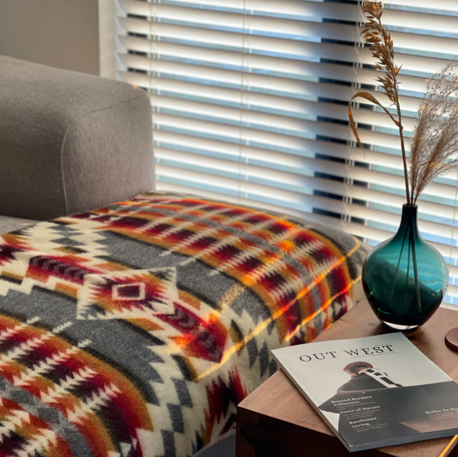 Patterned blanket on a sofa with a book titled 'Out West' and a vase on a table in the background.