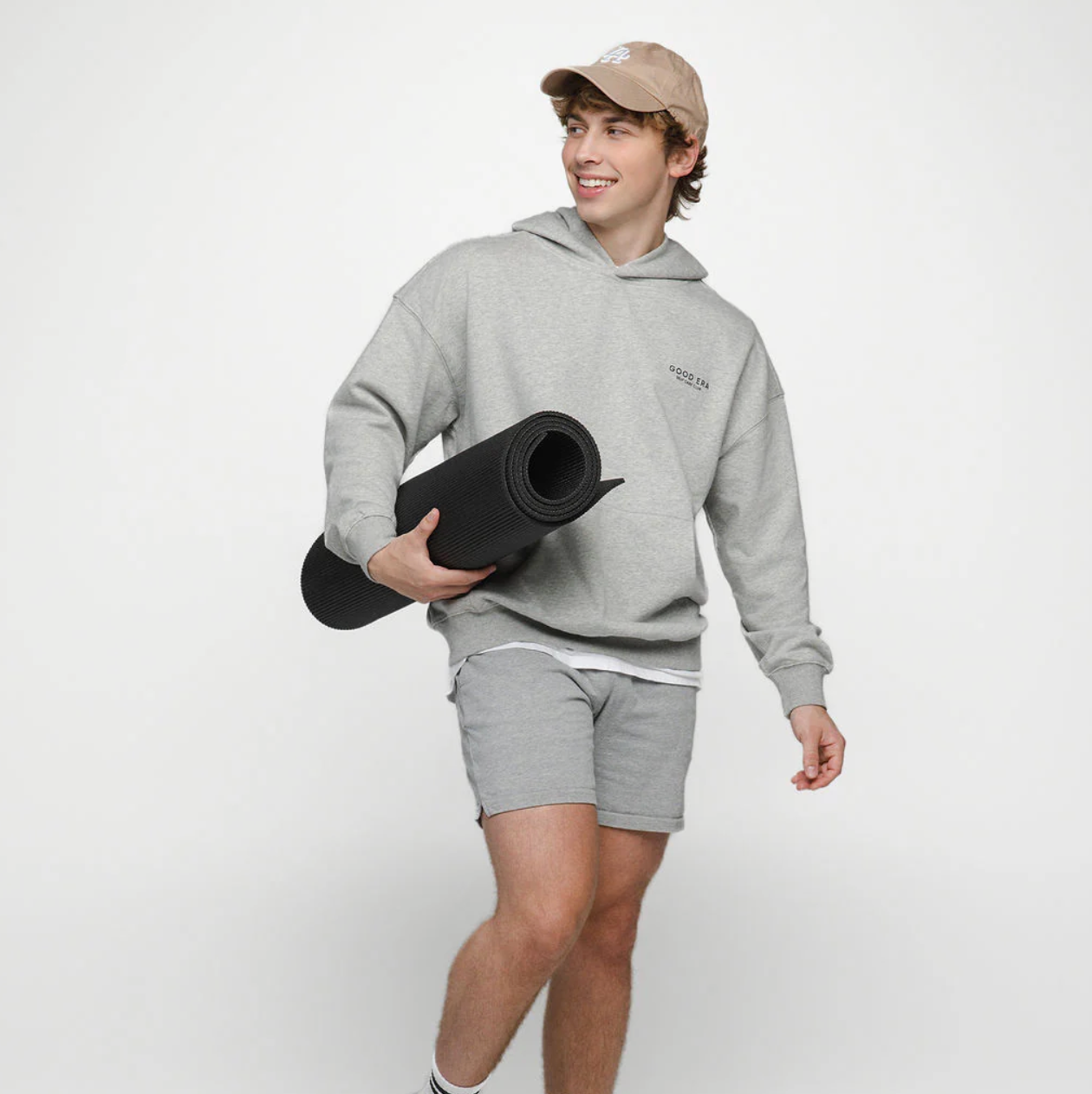 Person wearing a gray hoodie and shorts holding a black yoga mat on a white background