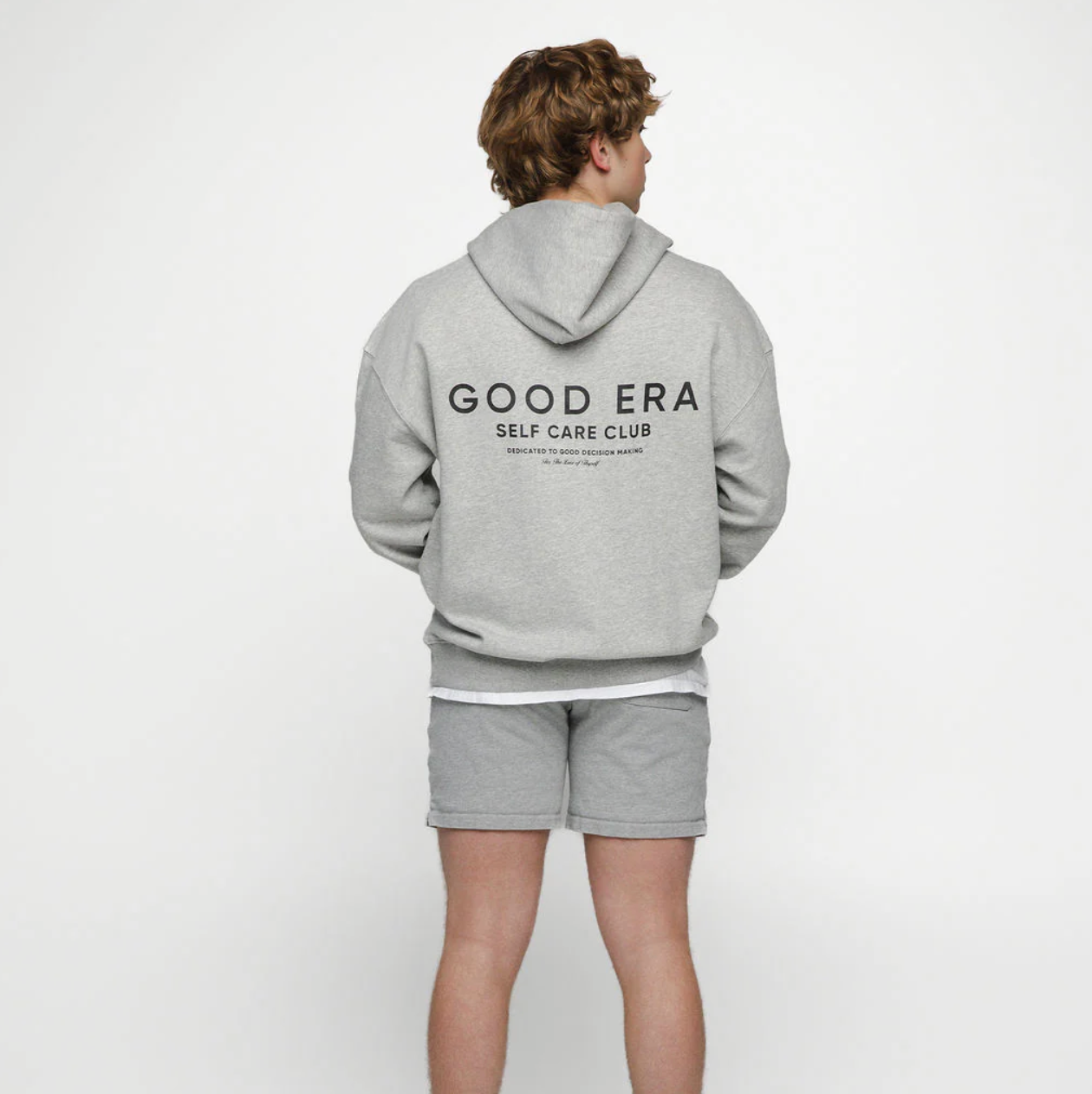 Person wearing a gray 'GOOD ERA Self Care Club' hoodie and shorts on a white background