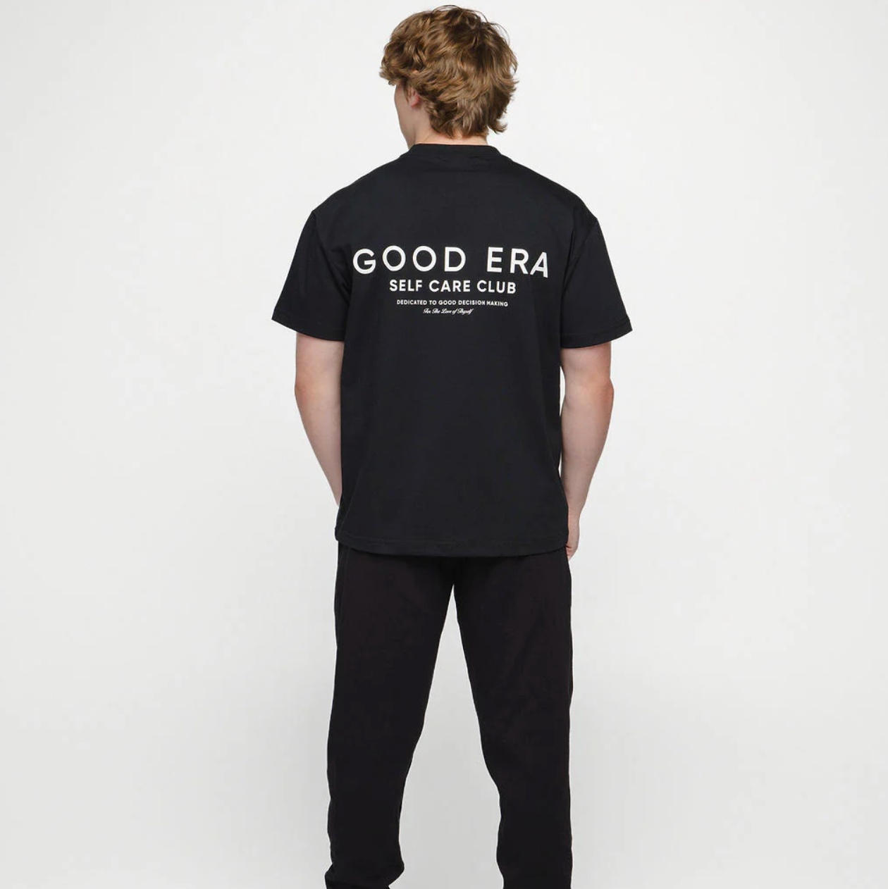 Person wearing a black t-shirt with 'GOOD ERA SELF CARE CLUB' text on the back against a white background