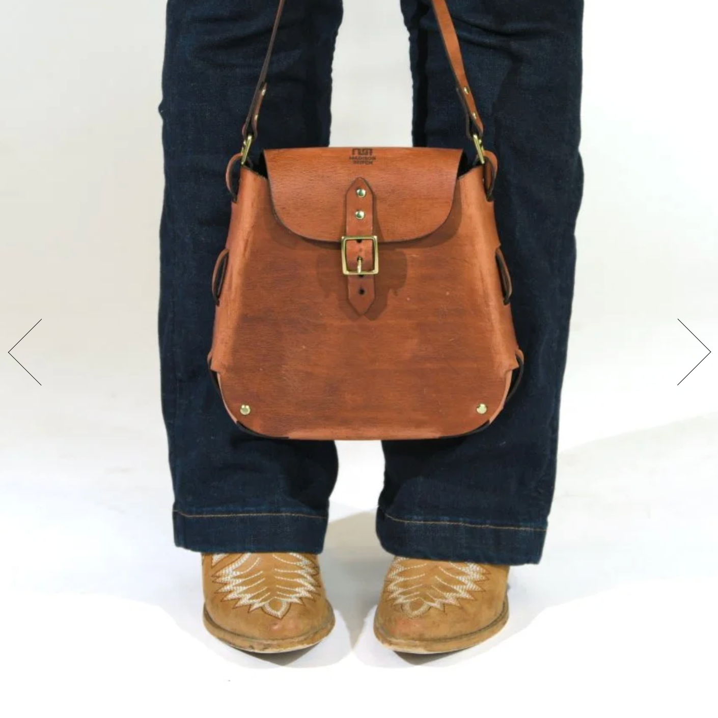 Brown leather bag worn over one shoulder with a person wearing jeans and brown shoes on a white background