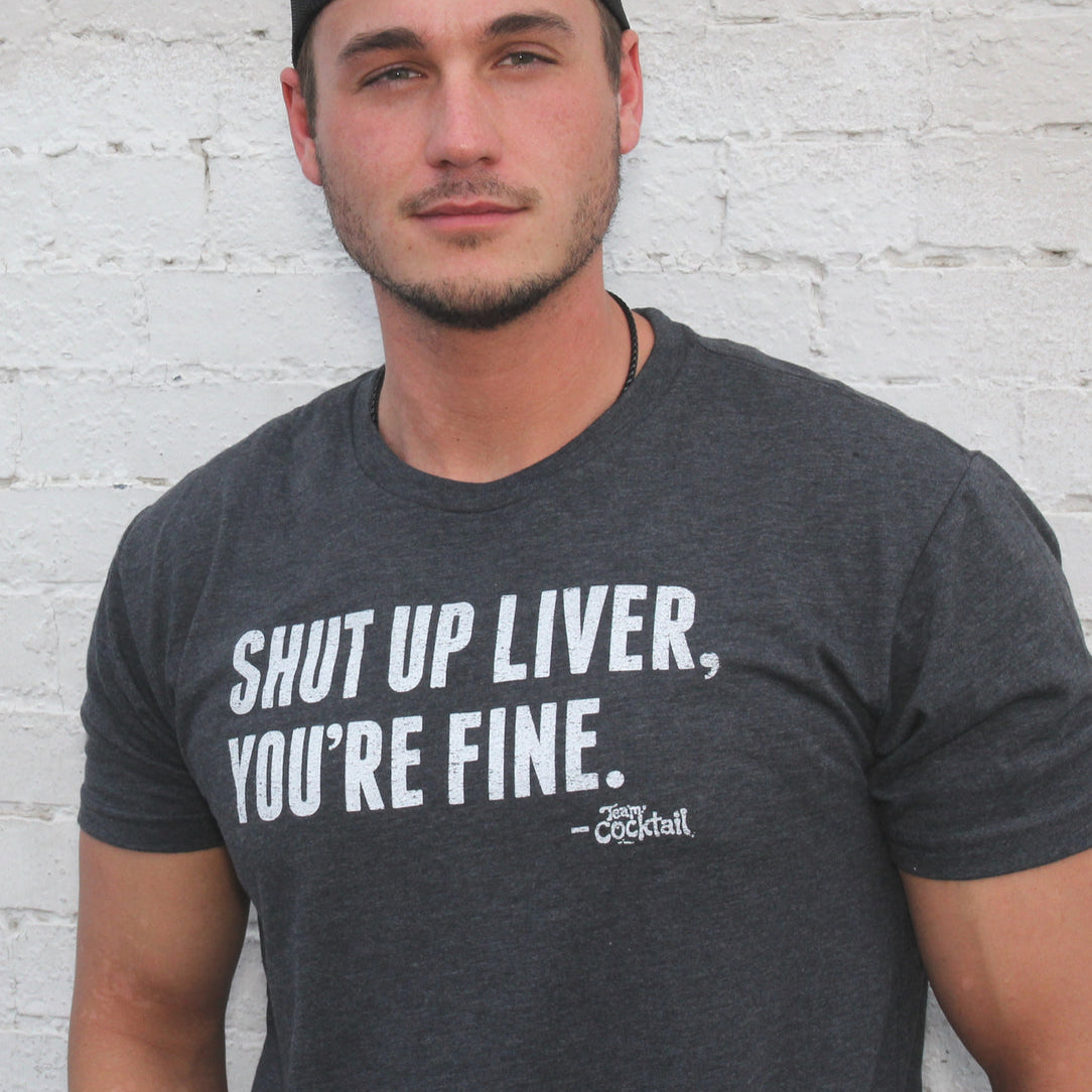 Man wearing a dark gray t-shirt with humorous text against a light brick wall.
