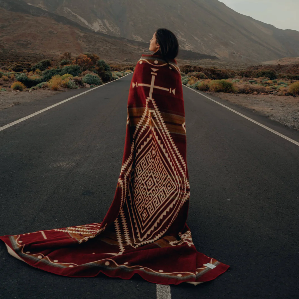 Person wrapped in a patterned blanket standing on a desert road with mountains in the background