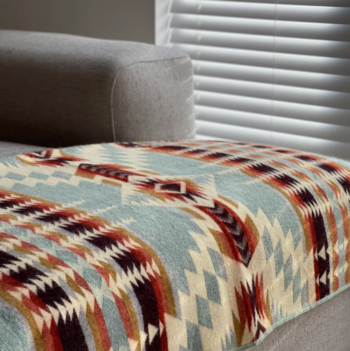 Patterned blanket draped over a gray sofa with blinds in the background