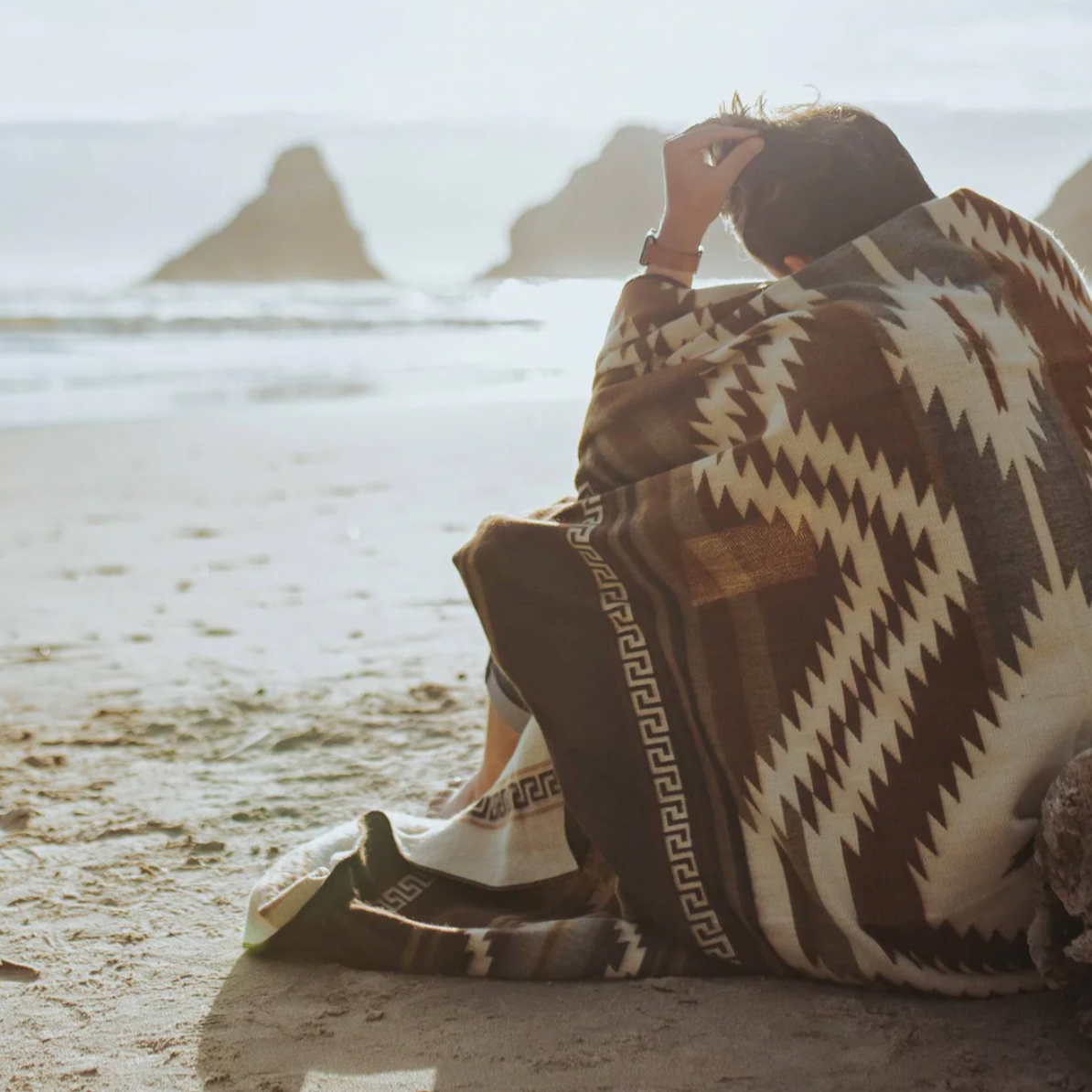 Person sitting on a sandy beach with a patterned blanket, wearing white sneakers.