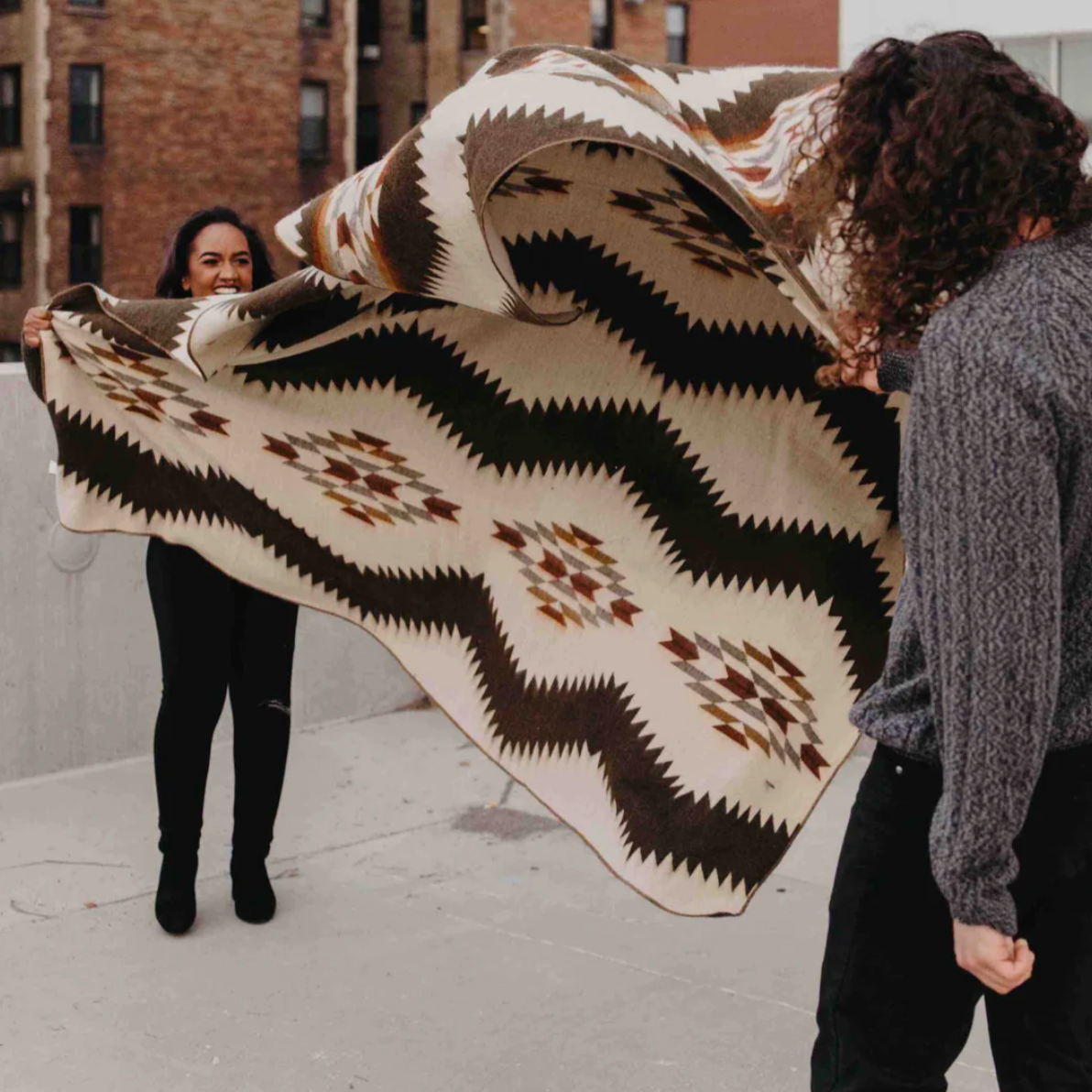 Two people holding a large patterned blanket outdoors.