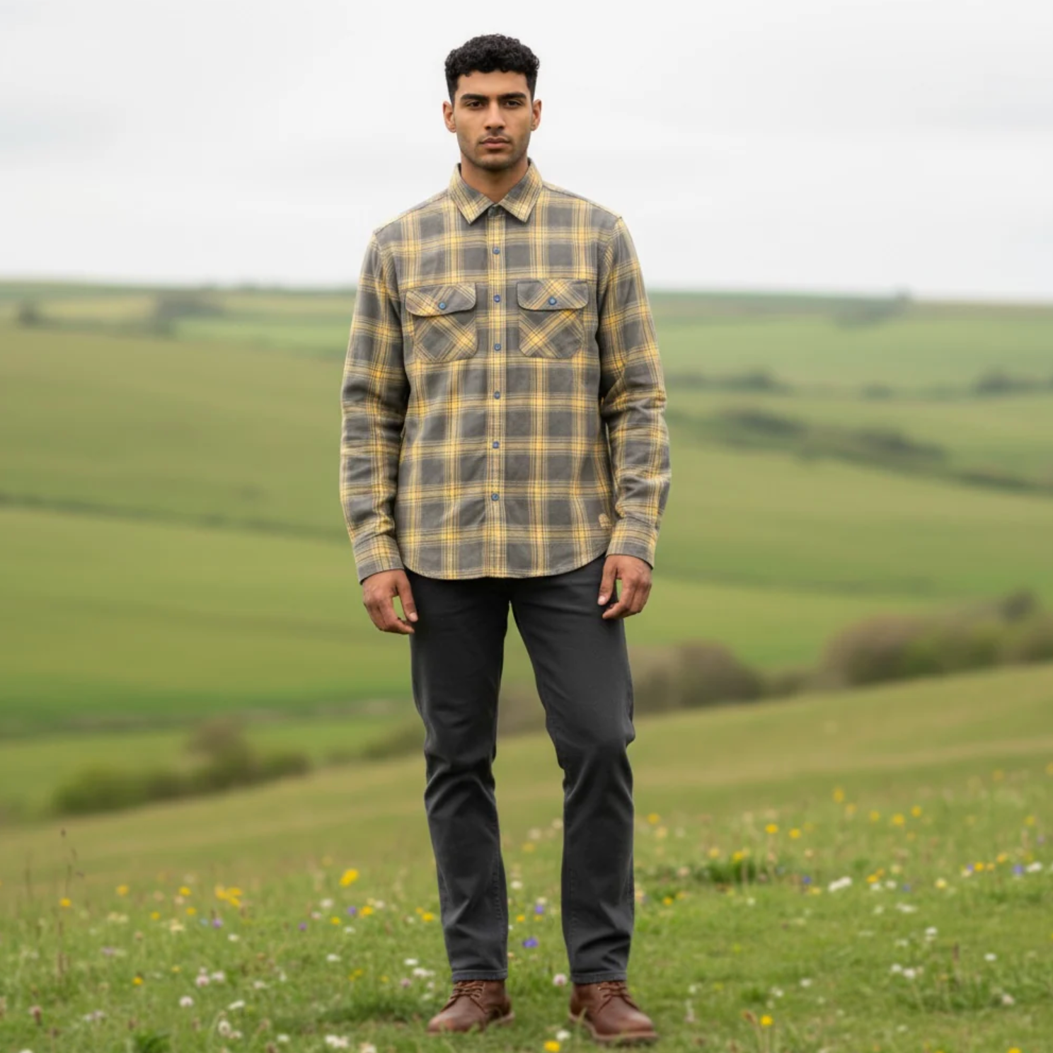 Man wearing a plaid shirt and dark pants standing in a field with green hills in the background